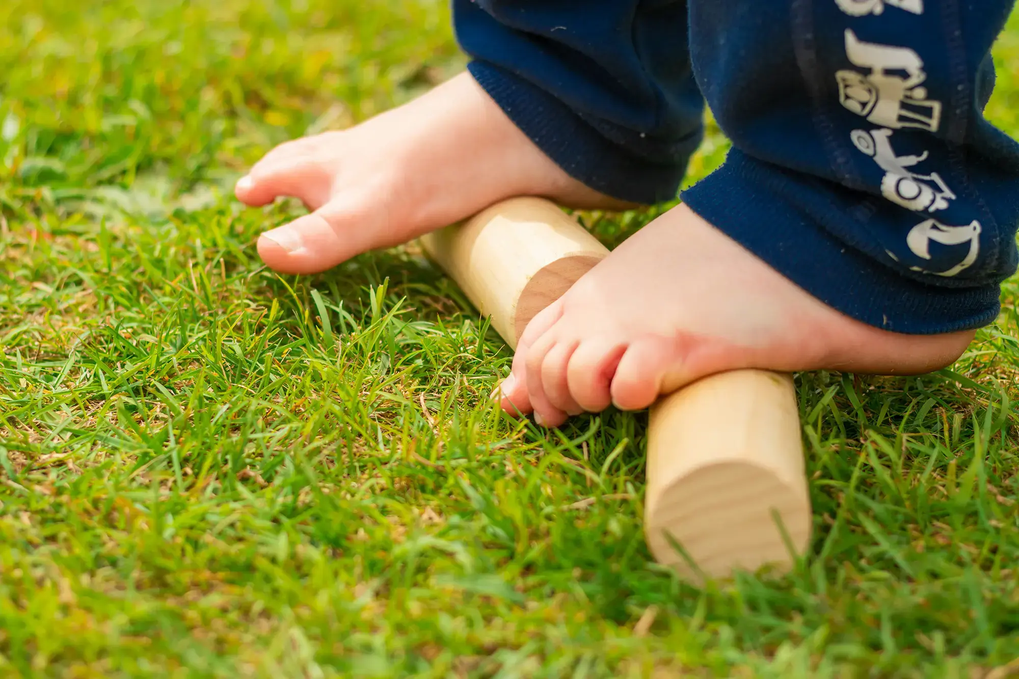 Kinderfüße balancieren auf runden Holzstäben in einer Wiese.